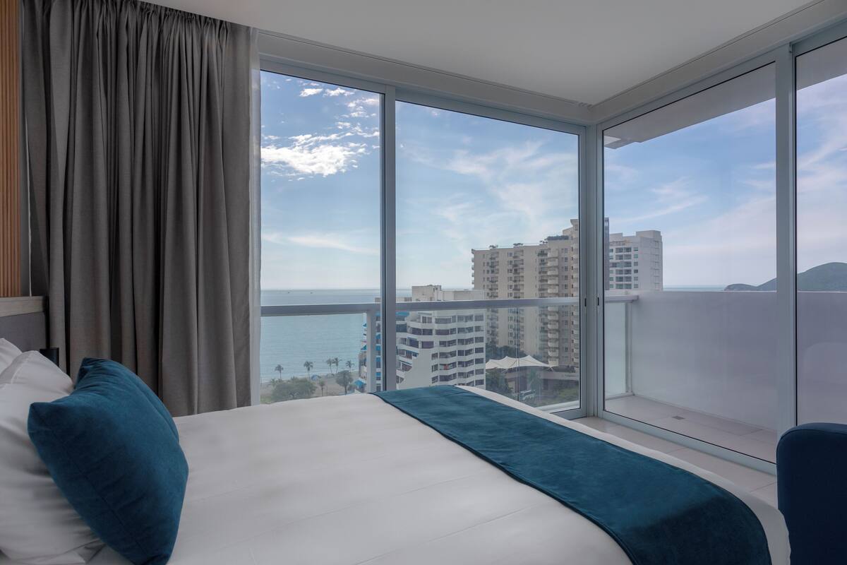 A hotel room with a large bed, a view of the sea, and high-rise buildings in the background.