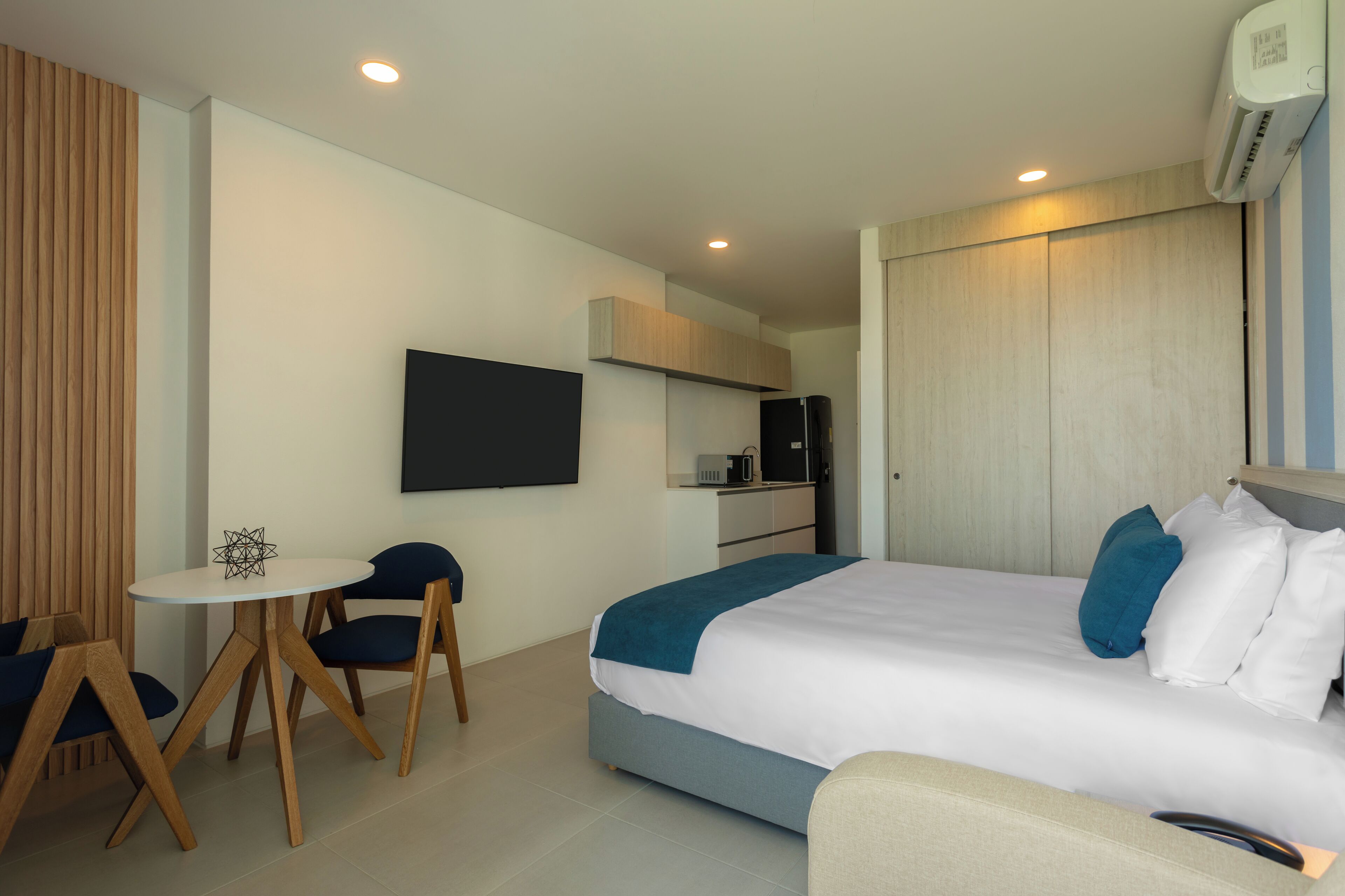 A hotel room with a bed, a desk with chairs, a television, a kitchenette, and a wardrobe.