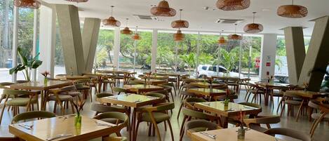 A spacious restaurant with multiple tables and chairs, large windows, and hanging wicker lamps.