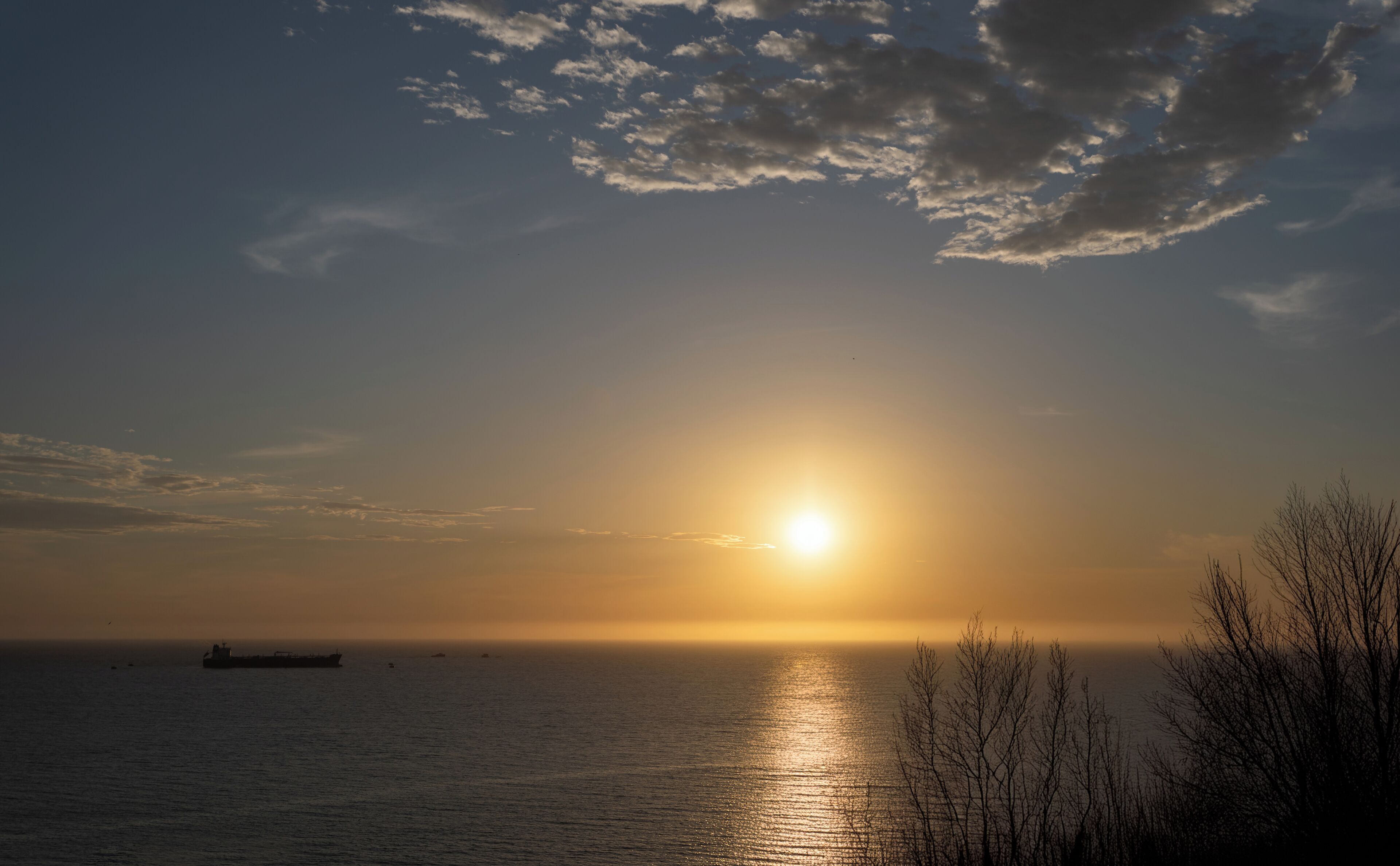 Ein Sonnenuntergang über ruhigem Meer, im Hintergrund ein Schiff und verschattete, kahlstehende Bäume vor dem Himmel.