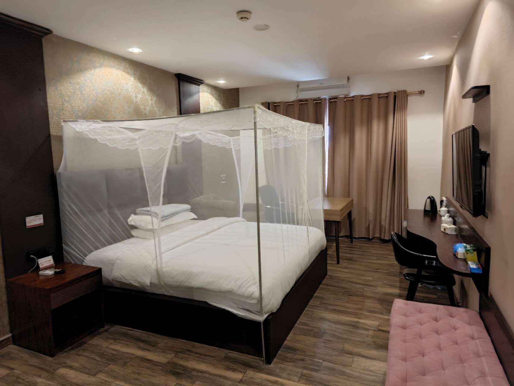 A hotel room with a four-poster bed, a desk, a chair, a bench, and a TV.