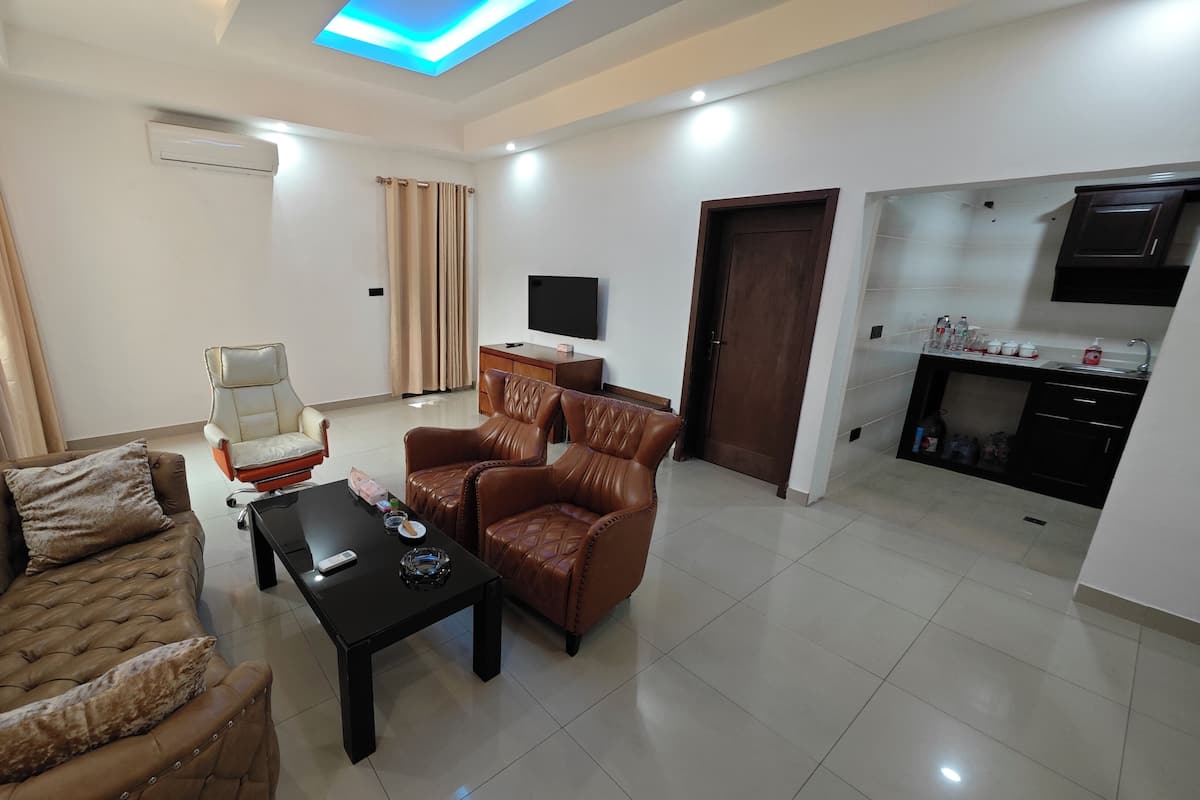 A living room with a sofa, armchairs, a coffee table, a TV, and a kitchen area with cabinets and a TV.