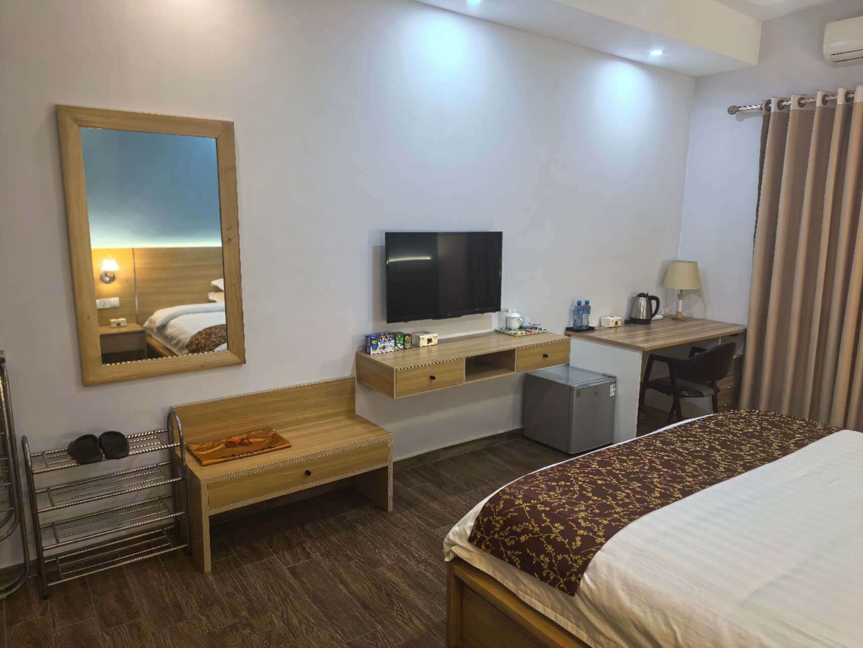 Habitación de hotel con cama, escritorio con silla, televisor, espejo, refrigerador pequeño y zapatero.