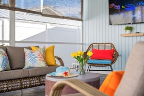 Habitación iluminada por el sol con un sofá, una silla de mimbre y una mesa redonda con un jarrón de tulipanes amarillos y un sombrero.