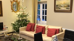 Un salon décoré d’un sapin de Noël, avec un canapé orné de coussins rouges, une table basse en verre et un tableau accroché au mur.