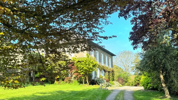 Un chemin mène à un grand bâtiment à la façade symétrique, entouré d’un espace vert bien entretenu et d’arbres.