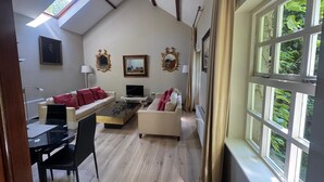 Un salon de style industriel avec un plafond en pente, un coin repas avec une table et des chaises, et une grande fenêtre donnant sur un paysage verdoyant.