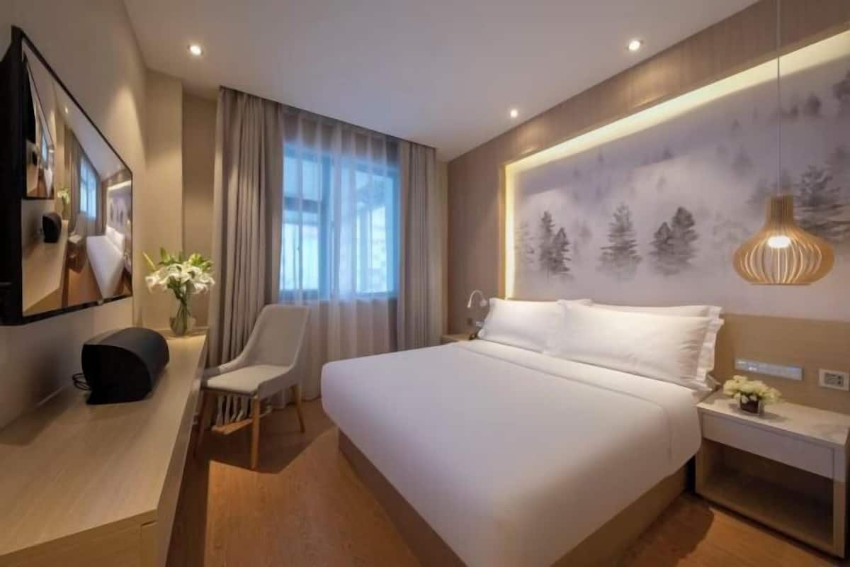 A modern hotel room with a large bed, a desk, a chair, a TV, and a wall-mounted artwork.