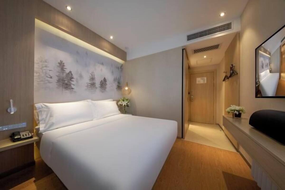 A modern hotel room with a large bed, a nightstand with a phone, a wall-mounted mirror, and a painting of trees.