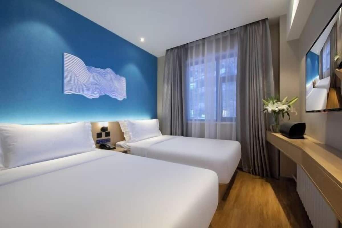 A hotel room with two beds, a large window with curtains, a wall-mounted artwork, and a bedside table with a lamp.