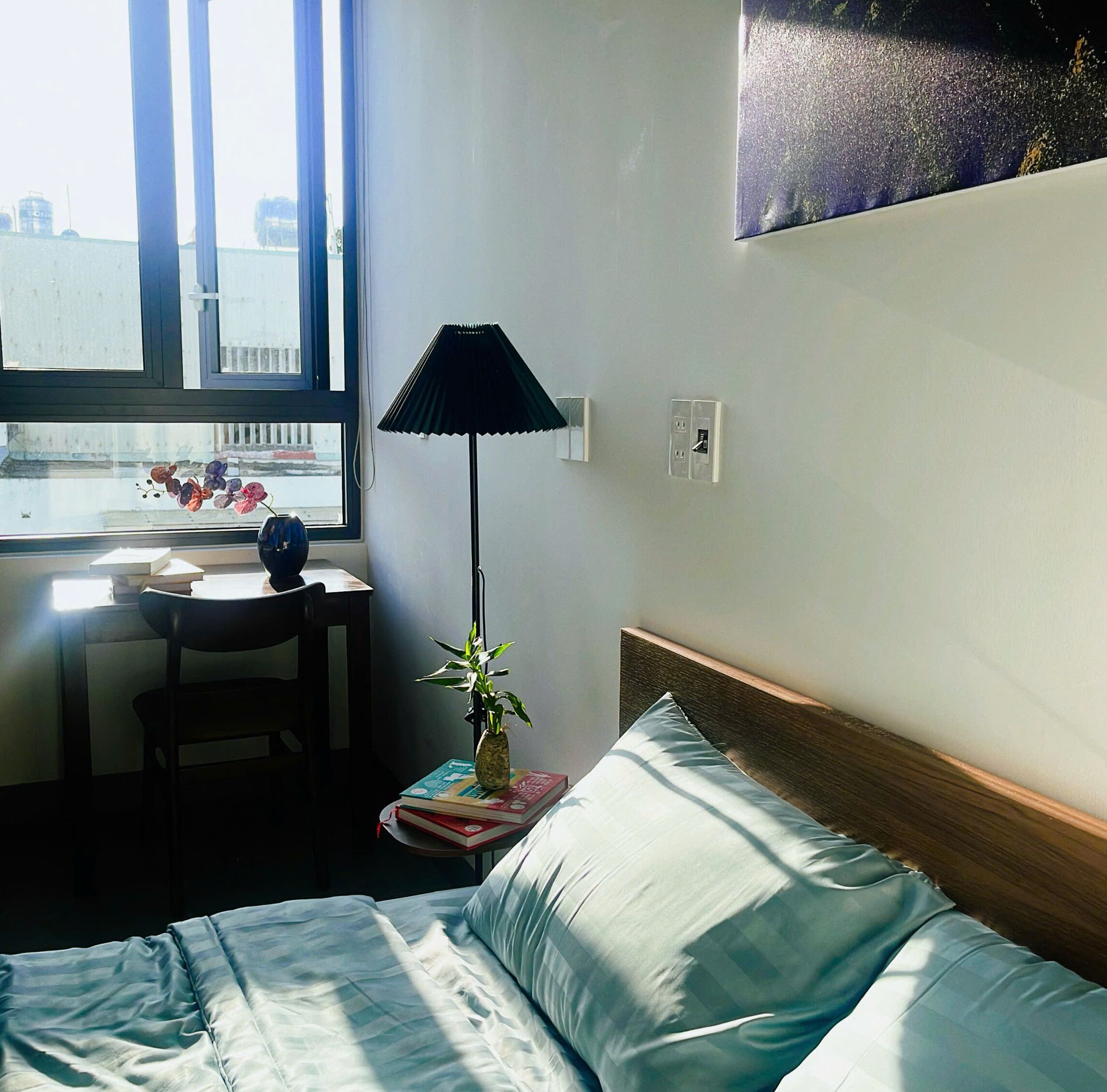 Habitación de hotel con cama, escritorio con silla, ventana y lámpara fijada a la pared.