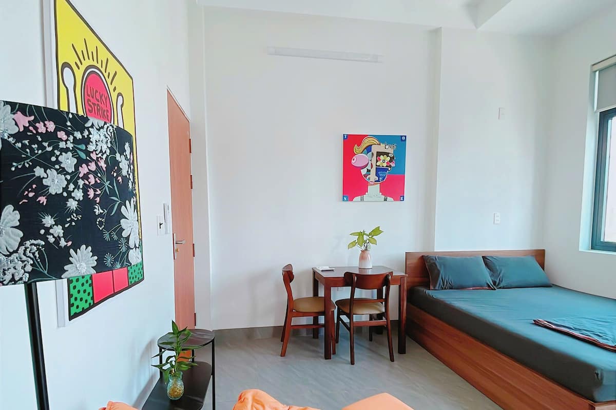 A small, modern room with a bed, a desk, a chair, and two paintings on the wall.