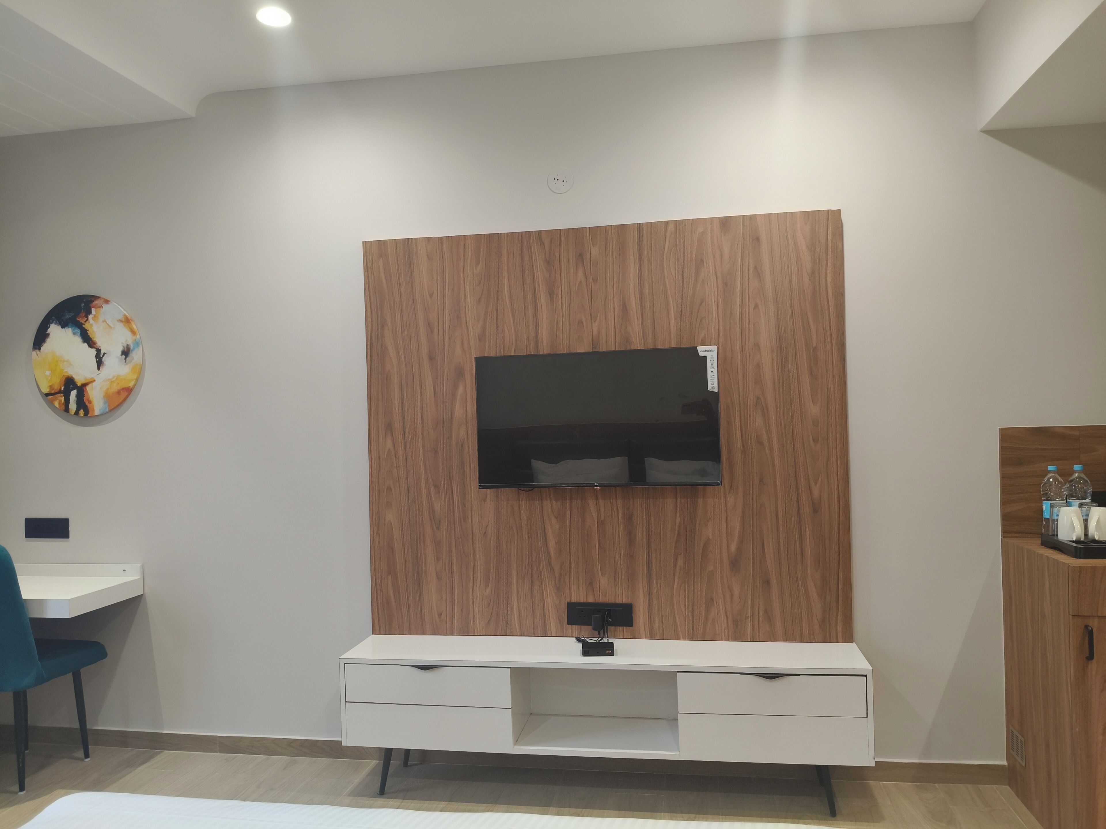 A modern living room with a flat-screen TV mounted on a wooden panel wall, a white entertainment unit below, a desk with a chair, and a colorful abstract painting on the wall.