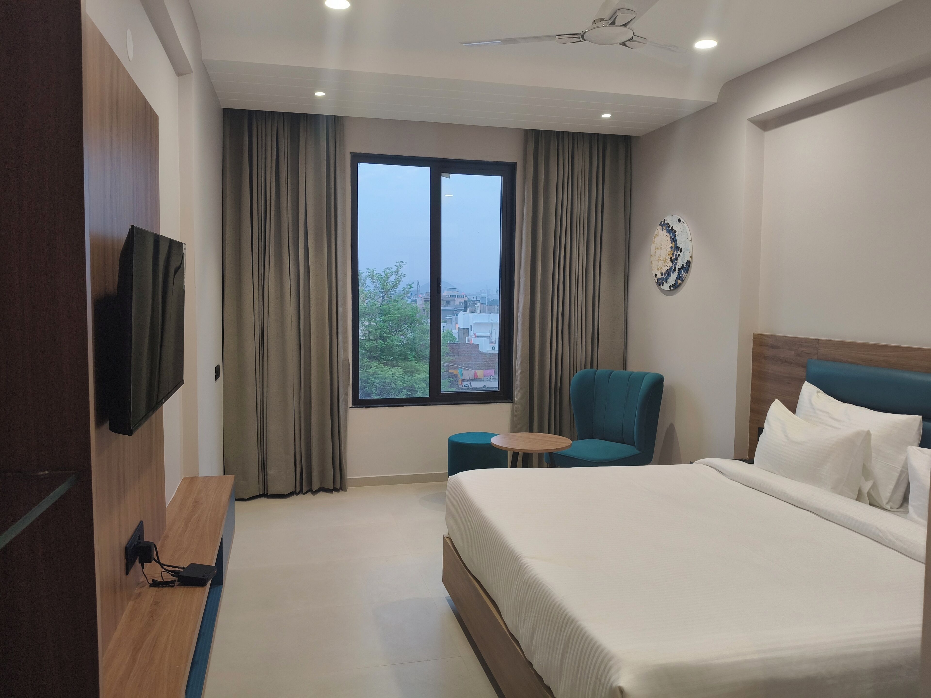 A hotel room with a bed, a chair, a small table, a TV, and a window with curtains.