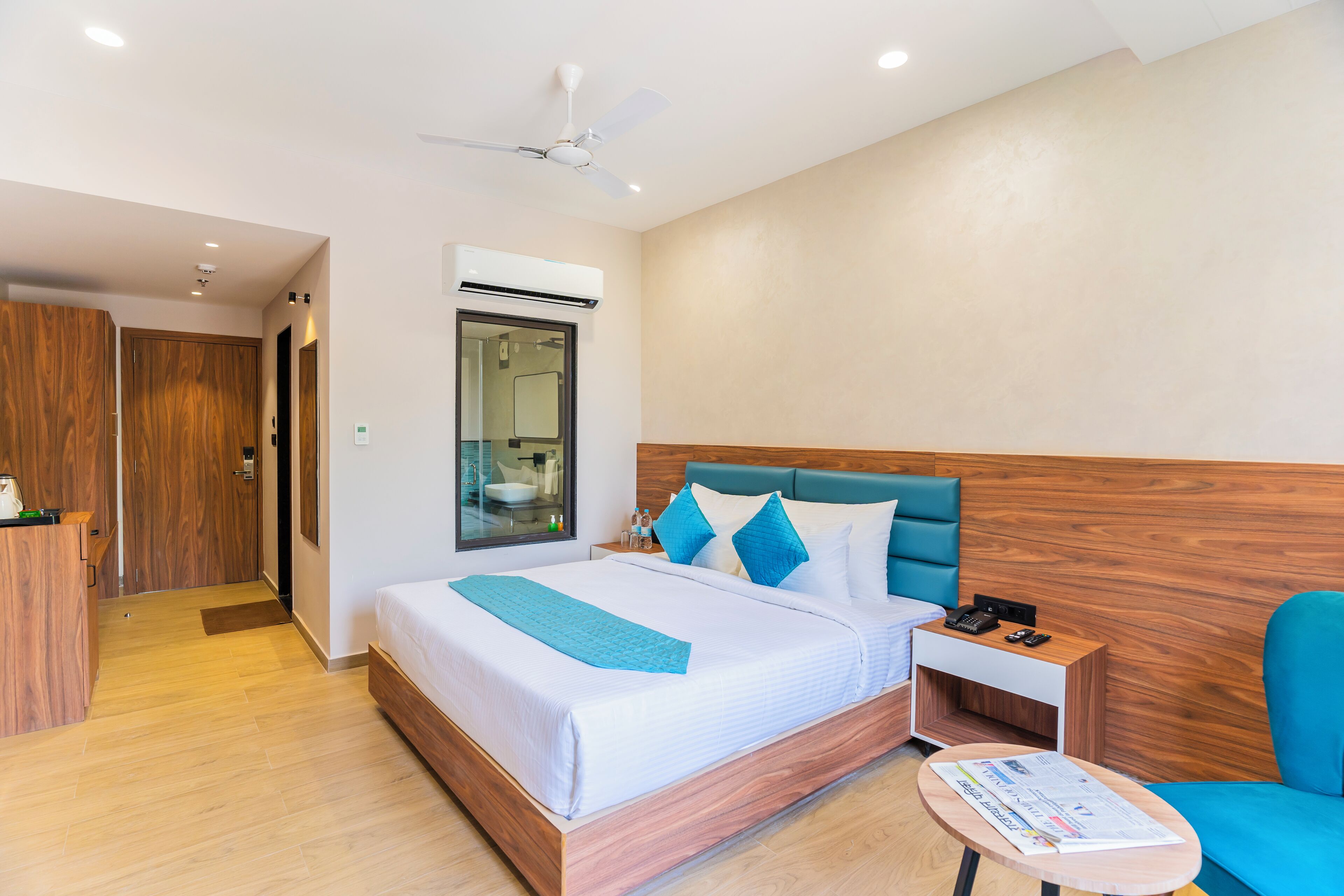 A modern hotel room with a large bed, a wooden headboard, a nightstand, a blue chair, and a wooden wardrobe.