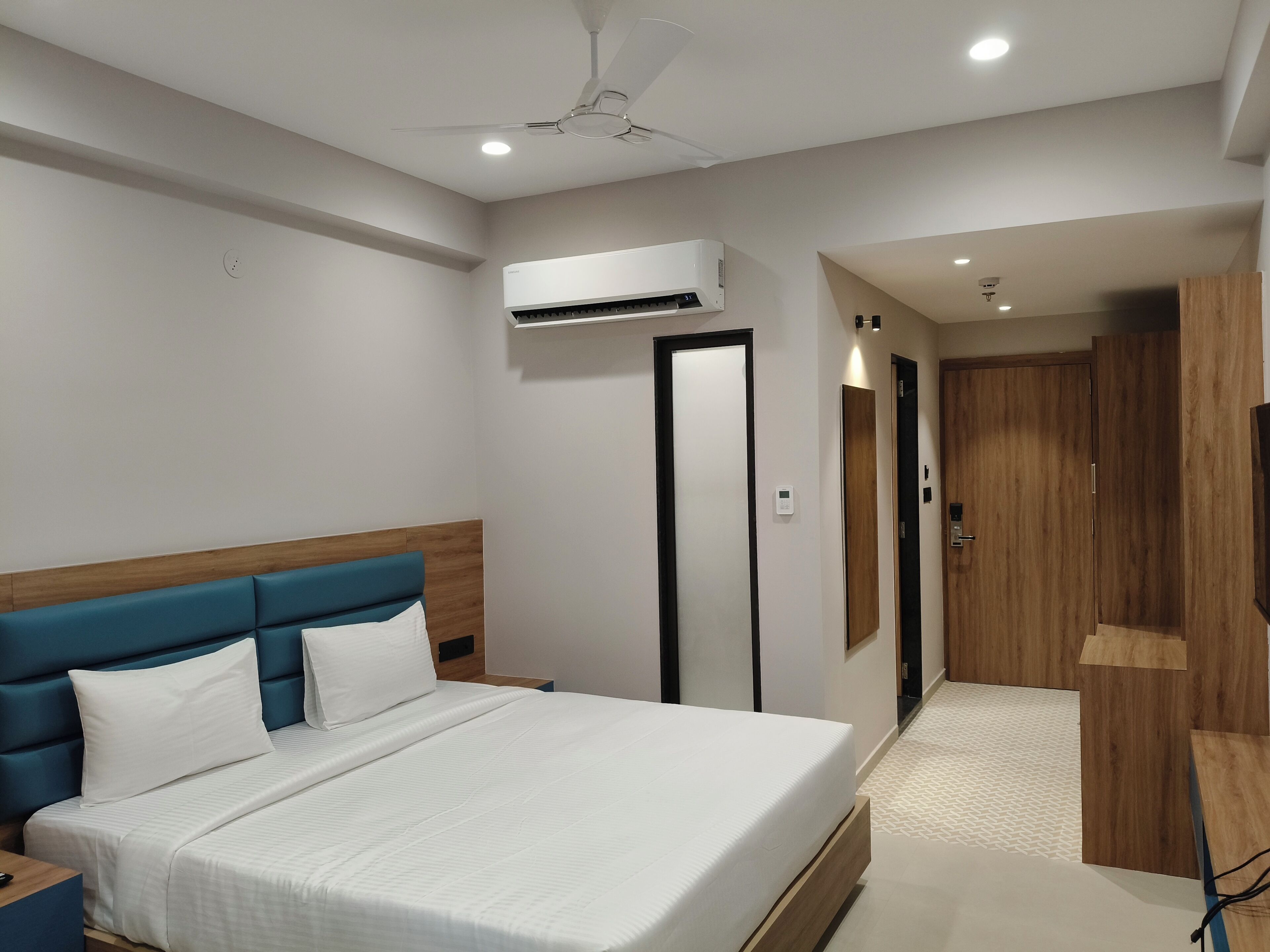 A modern hotel room with a large bed, a nightstand, a ceiling fan, an air conditioning unit, and a wooden wardrobe.