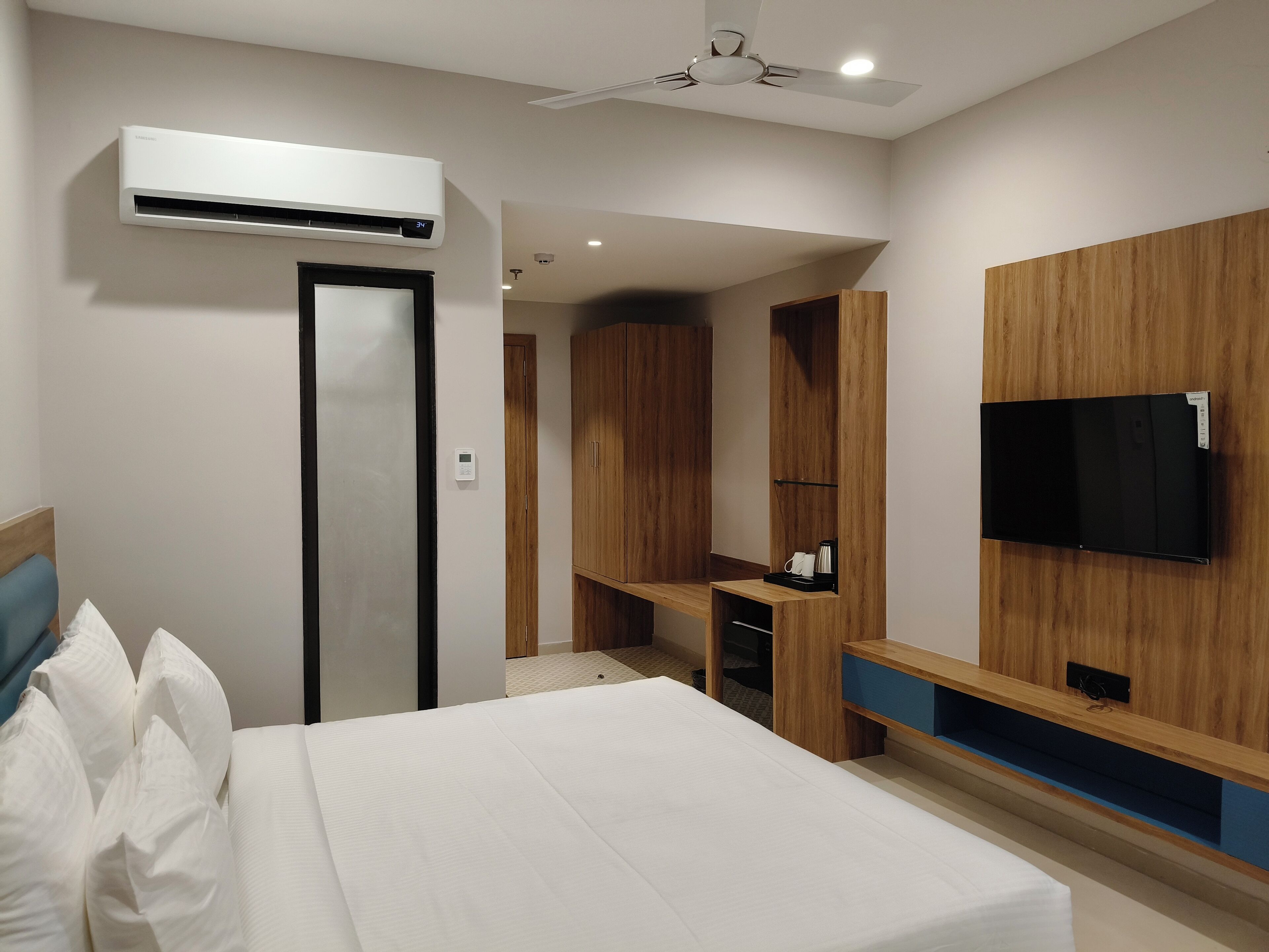 A modern hotel room with a bed, a TV mounted on the wall, a wooden wardrobe, and an air conditioning unit.