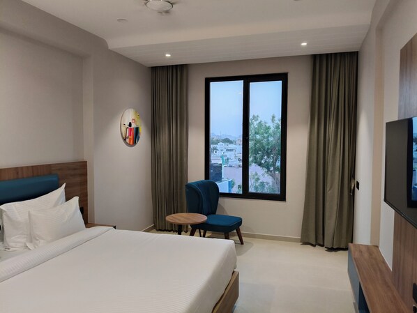 A hotel room with a large bed, a small round table, a blue chair, a TV, and a window with curtains.