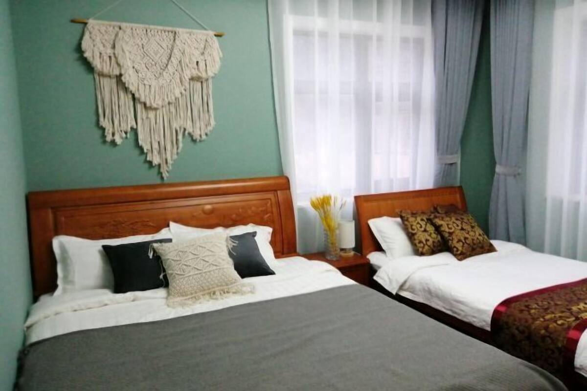 A hotel room with two beds, a wooden headboard, a macrame wall hanging, a vase with flowers, and a window with curtains.