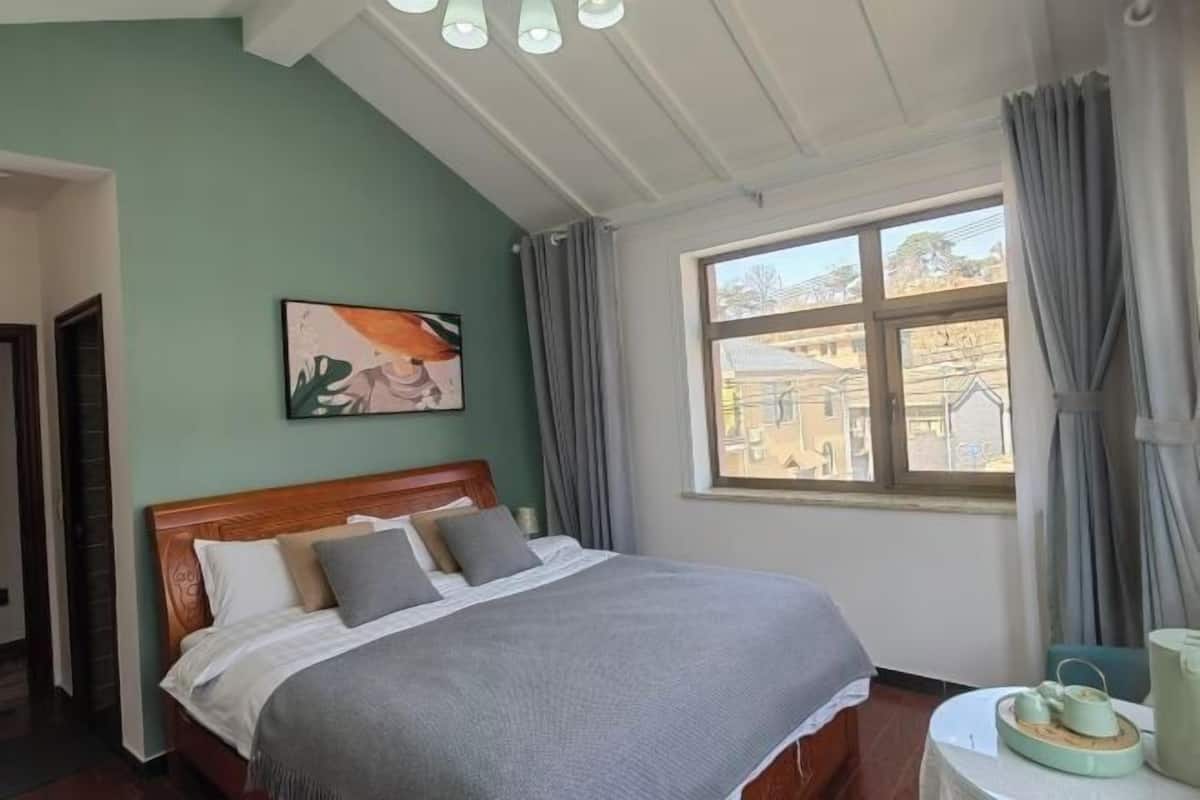 A neatly made bed with a wooden headboard, a painting on the wall, and a window with curtains.