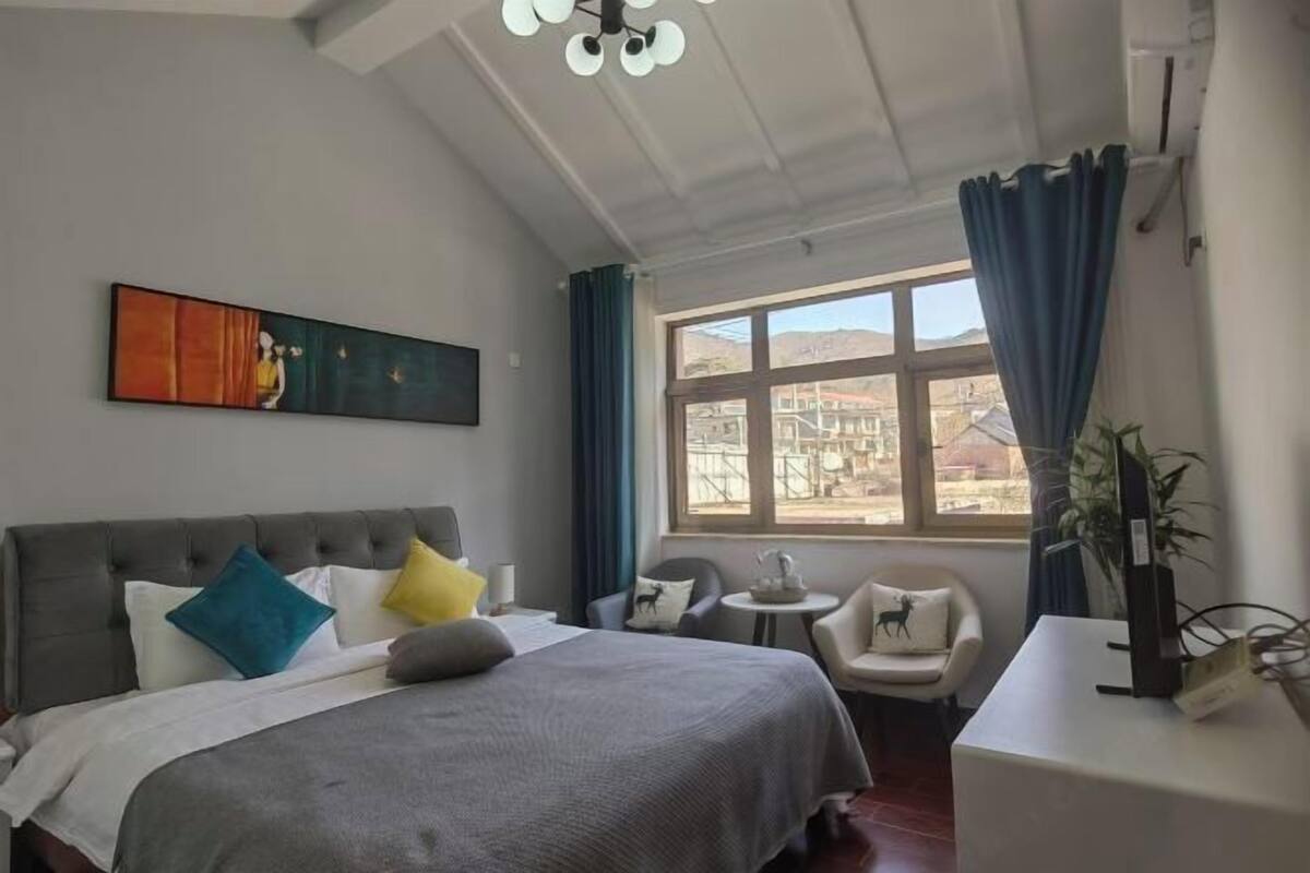 A hotel room with a bed, a desk, a chair, a window with curtains, a painting on the wall, and a ceiling fan.