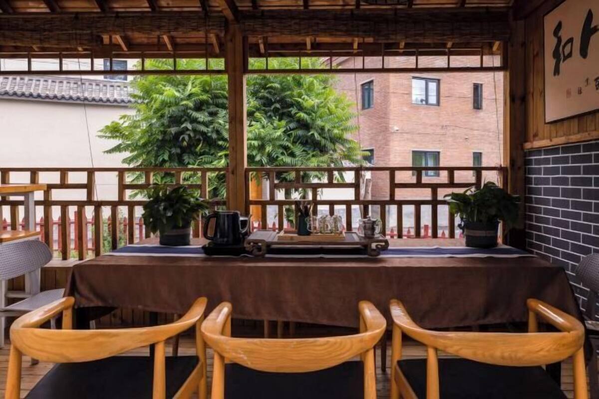 A wooden deck with a table set for dining, surrounded by chairs, and a view of a brick building and greenery.