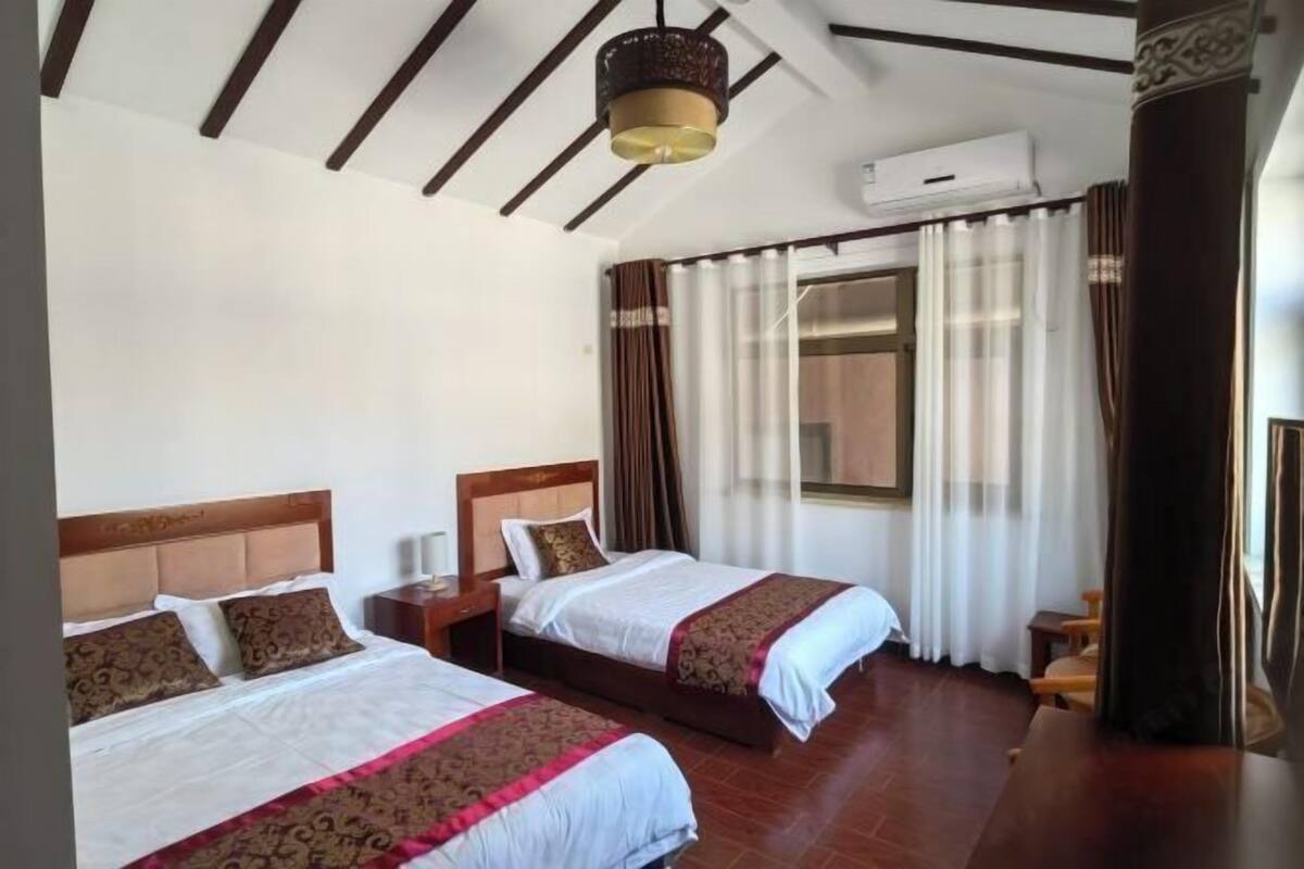 A hotel room with two beds, a wooden ceiling, an air conditioning unit, and a window with curtains.