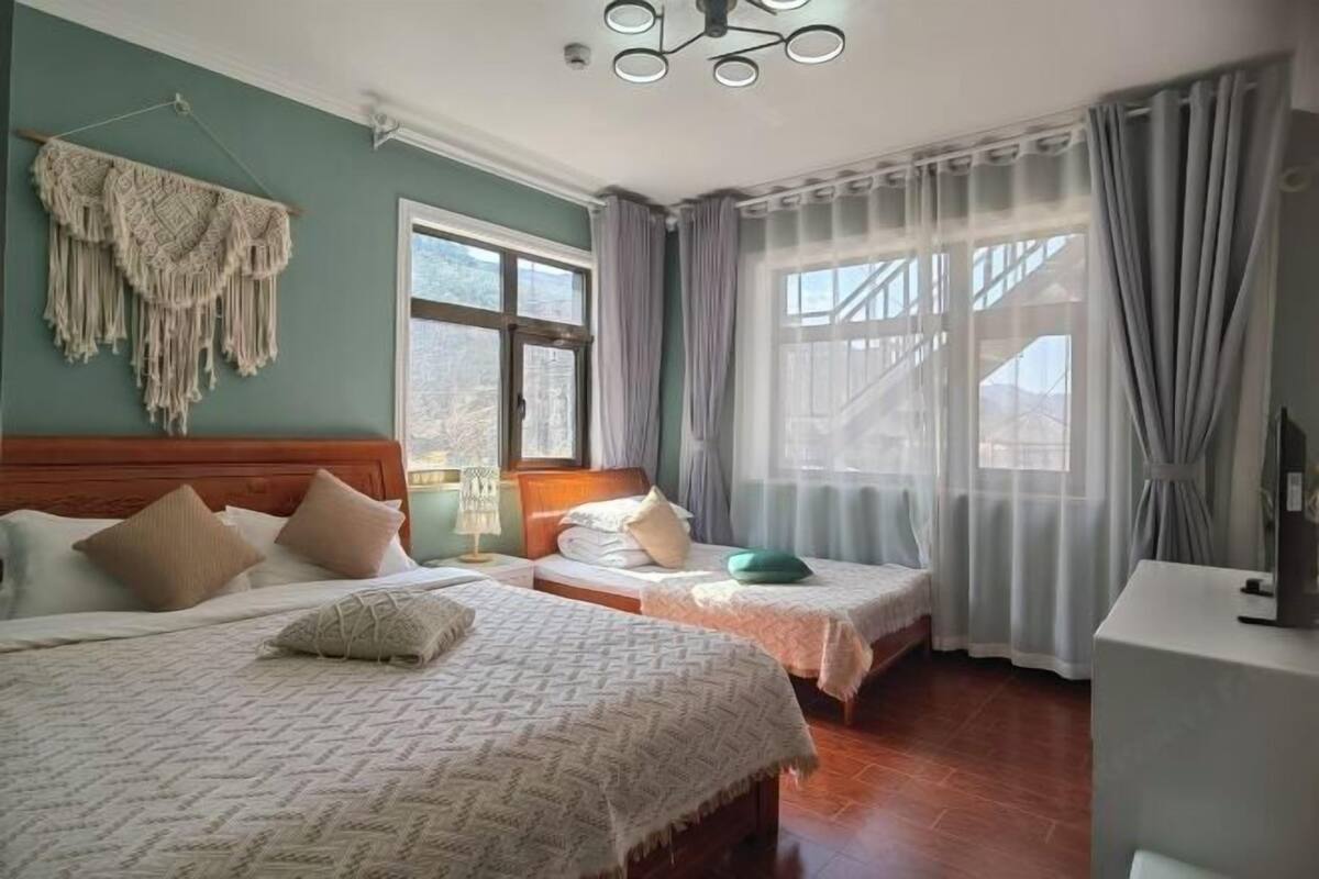 A hotel room with two beds, a wooden headboard, a large window with curtains, and a macrame wall hanging.