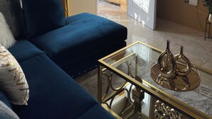 Ein Wohnzimmer mit einem blauen Sofa, einem Glastisch mit Dekoration und Blick durch eine offene Tür auf einen Außenbereich mit Geländer und Grünpflanzen.
