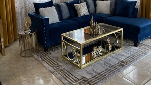 Ein Wohnzimmer mit einem blauen Ecksofa, einem Glastisch und gemusterten Vorhängen.