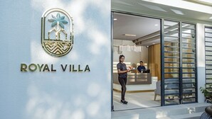 Exterior de un hotel llamado “ROYAL VILLA” con un diseño moderno, que incluye una entrada de vidrio y se puede ver el interior con asientos y un escritorio.