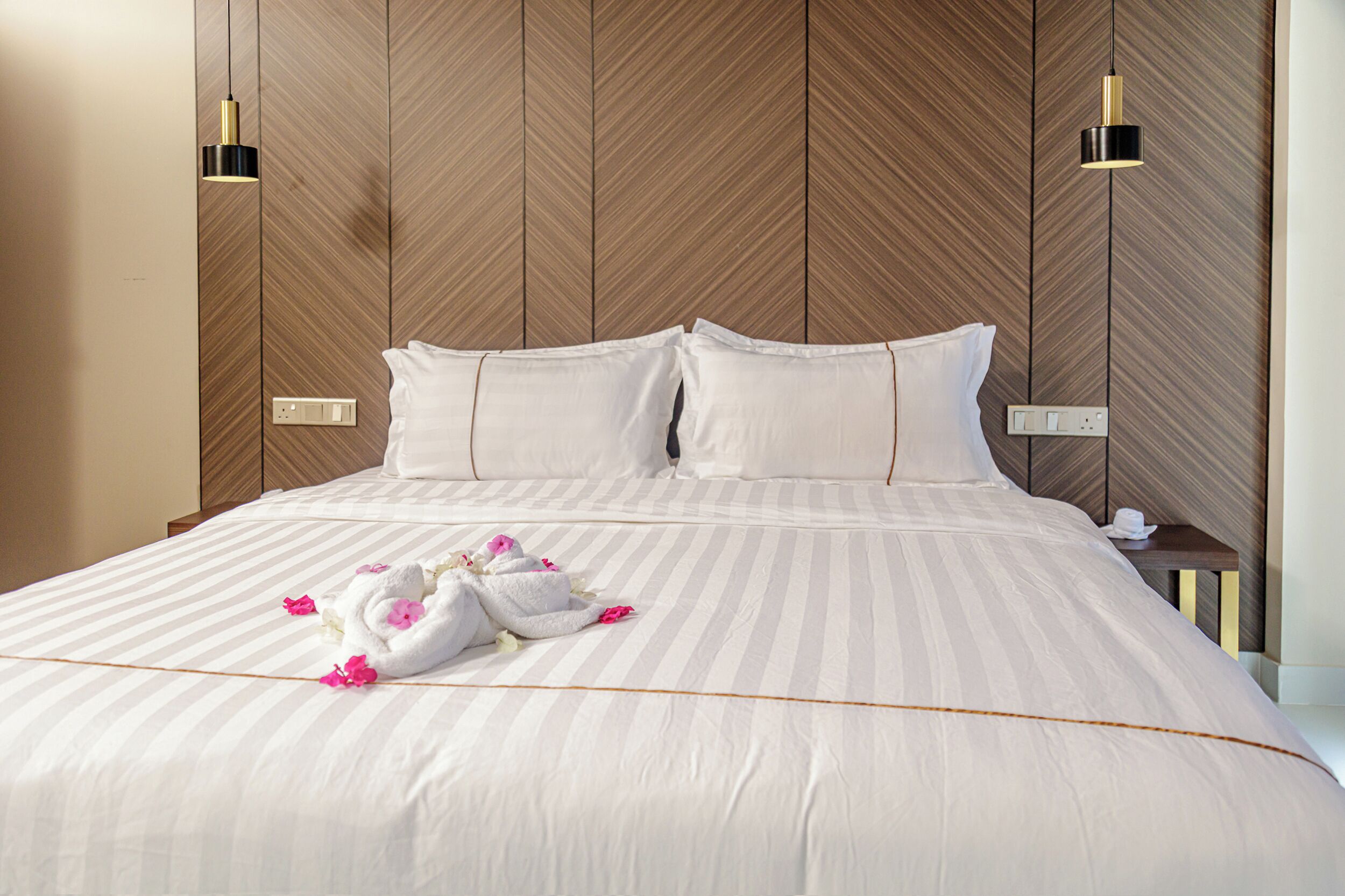 Una cama bien tendida con sábanas blancas y flores rosadas, un cabecero de madera y dos lámparas de noche.