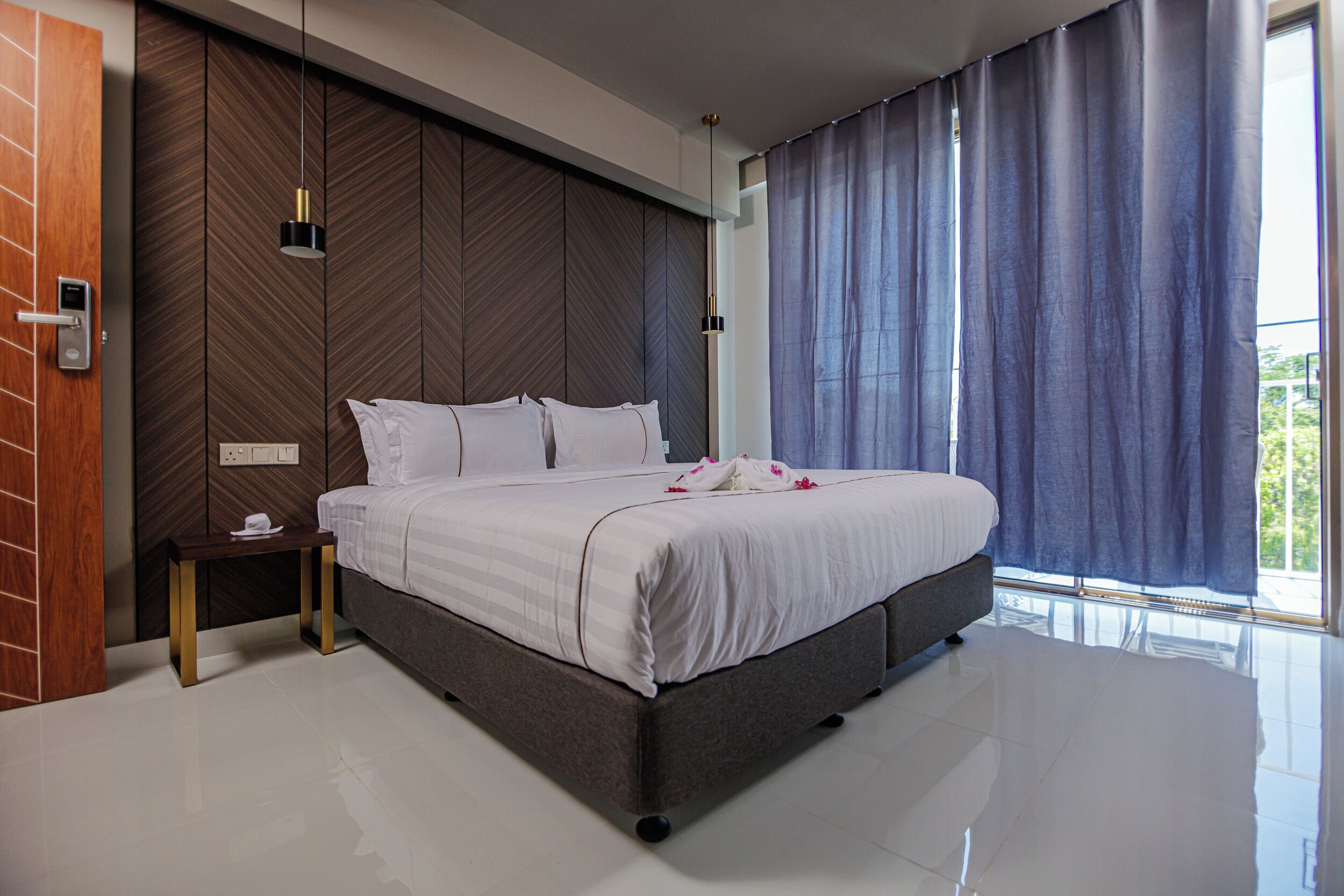 A modern hotel room with a large bed, a wooden accent wall, a small bedside table, and a large window with curtains.
