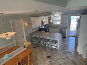 Cozinha moderna com armários brancos, uma ilha central com banquetas e um espaço de jantar com mesa e cadeiras de madeira.