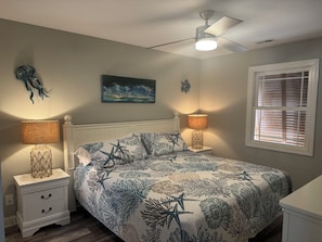 Quarto com cama, mesas de cabeceira, ventoinha no teto, um quadro e uma janela com persianas.