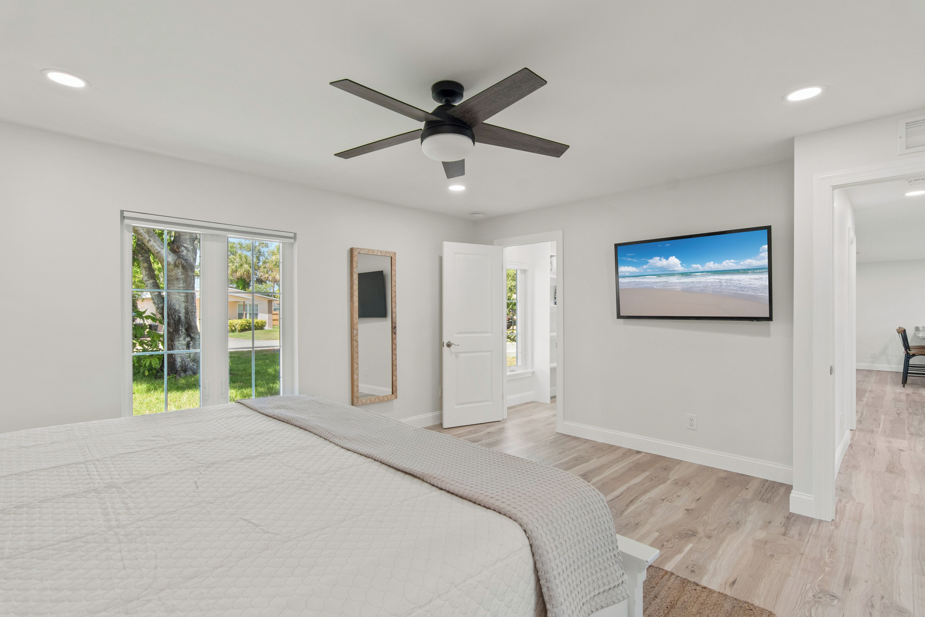 Une chambre à coucher comprenant un lit, un ventilateur de plafond, une grande fenêtre et une porte donnant sur une autre pièce.