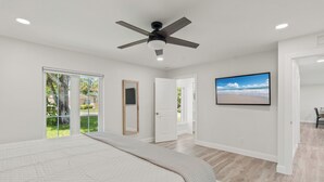 Une chambre à coucher comprenant un lit, un ventilateur de plafond, une grande fenêtre et une porte donnant sur une autre pièce.