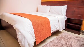 Una cama bien hecha con ropa de cama blanca y naranja, un cabecero de madera y una alfombra rosa en el suelo.