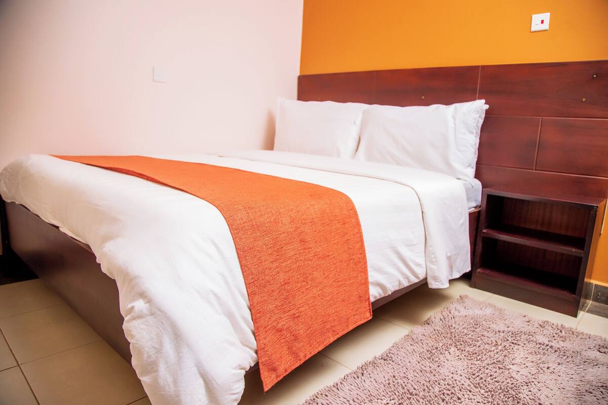 Ein ordentlich bezogenes Bett mit weißer und orangefarbener Bettwäsche, einem hölzernen Kopfteil und einem pinkfarbenen Teppich auf dem Boden.