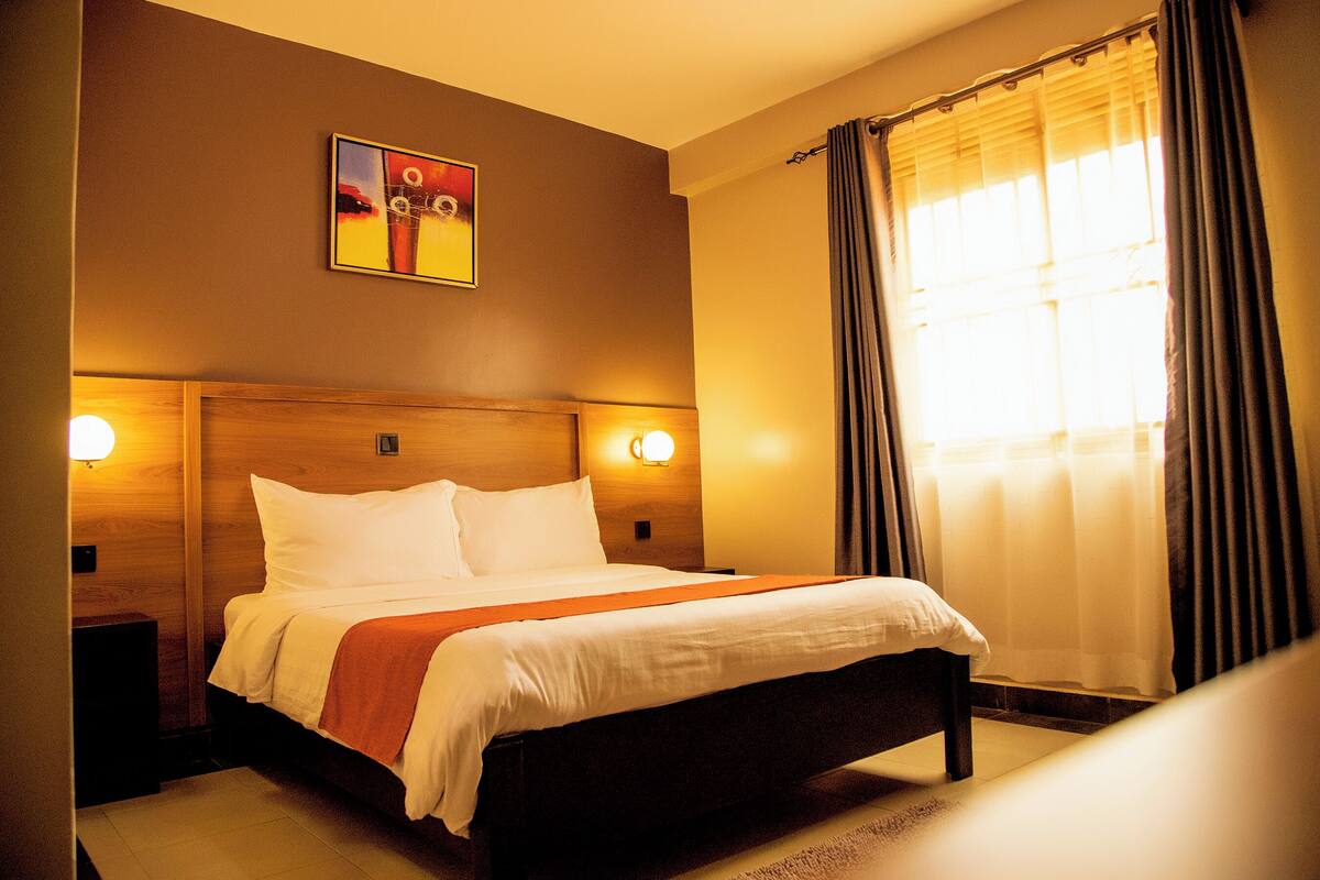 Ein Hotelzimmer mit einem großen Bett, zwei Nachttischen, einer Kopfteil und einem Gemälde an der Wand, sowie einem Fenster mit Vorhängen.