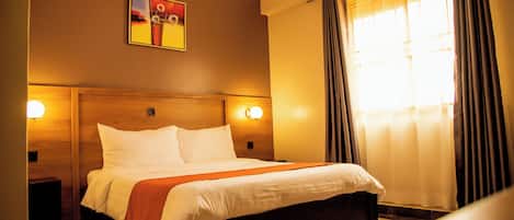 A hotel room with a large bed, two bedside tables, a headboard, a painting on the wall, and a window with curtains.