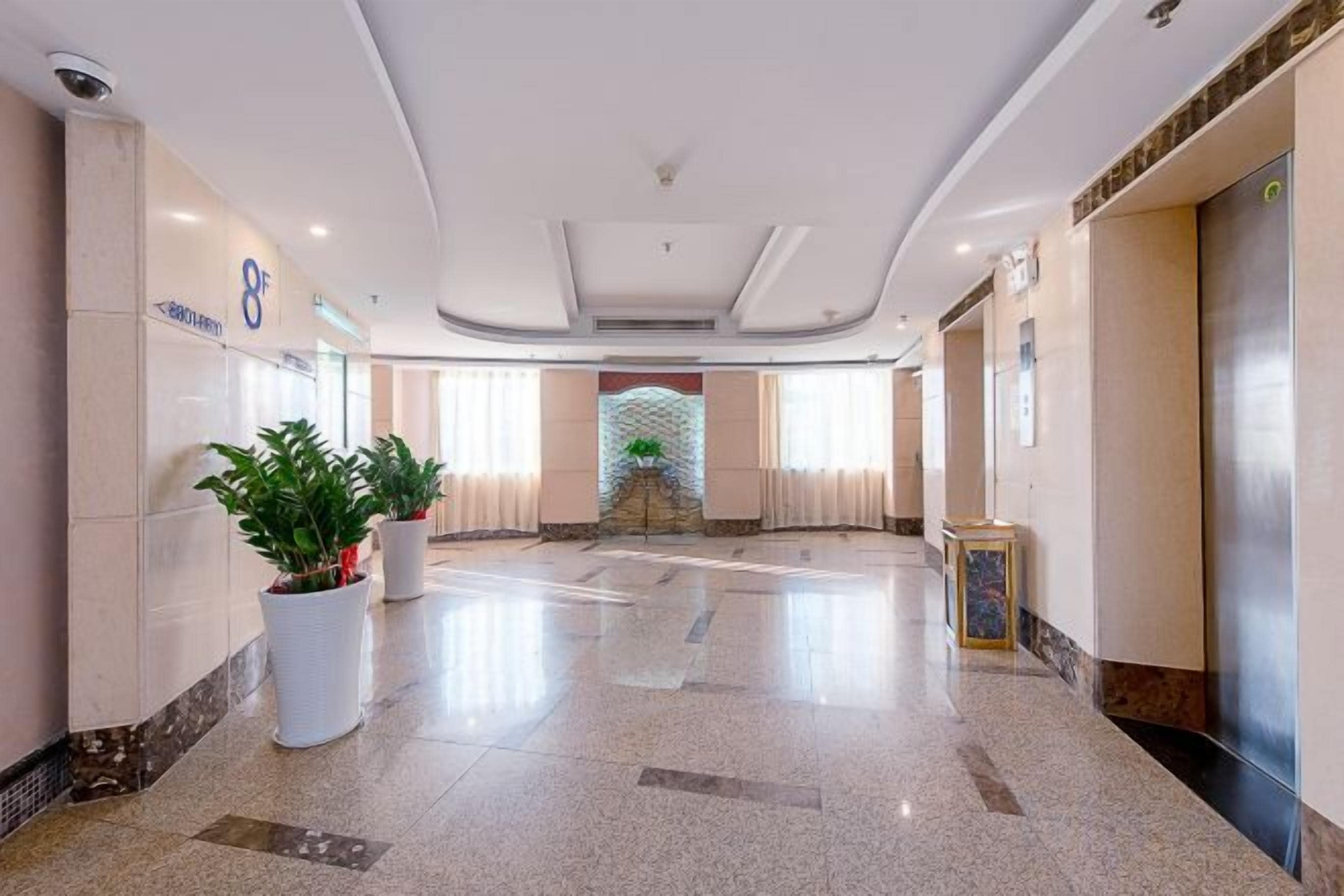 A hotel lobby with a reception desk, potted plants, a large mirror, and an elevator.