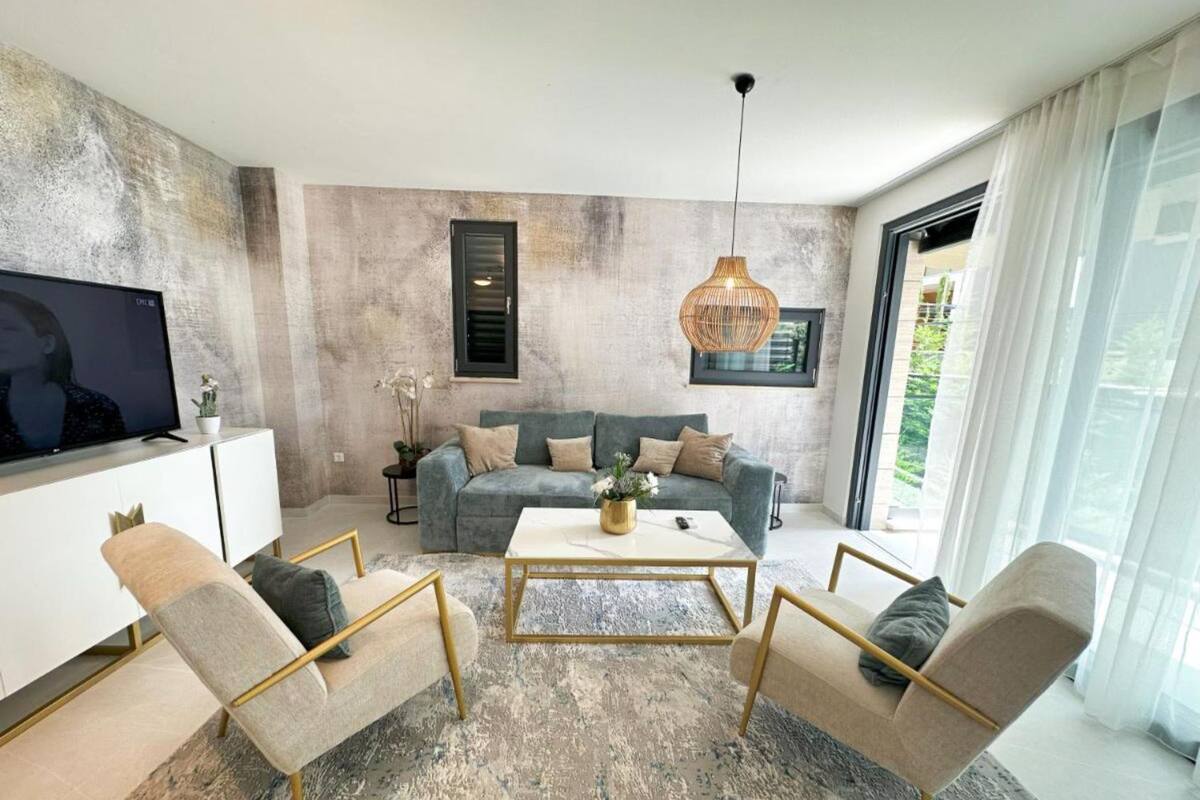 A modern living room with a grey sofa, a coffee table, and two armchairs. There is a TV on a white cabinet, a hanging light fixture, and a window with curtains.