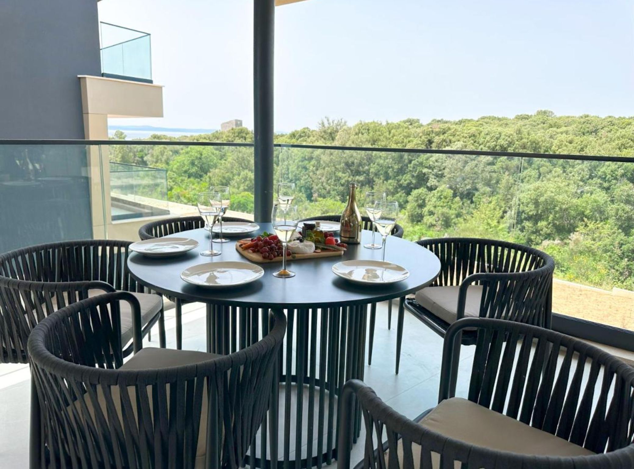A round table with a glass top, surrounded by black wicker chairs, set for a meal with plates, glasses, and a bottle of wine.
