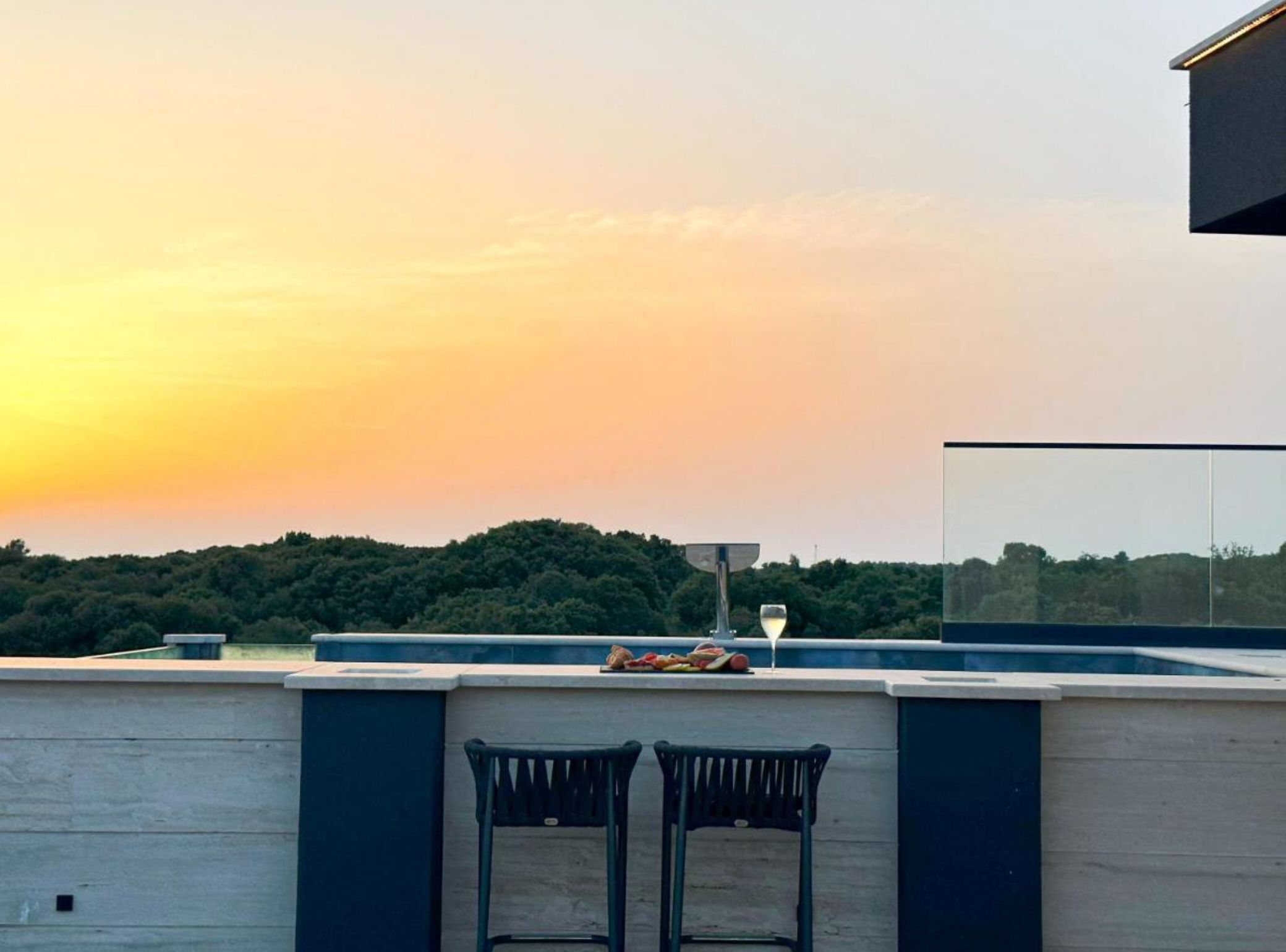 Une terrasse sur le toit avec deux chaises, une table garnie de nourriture et un verre de vin, offrant une vue sur une zone boisée au coucher du soleil.