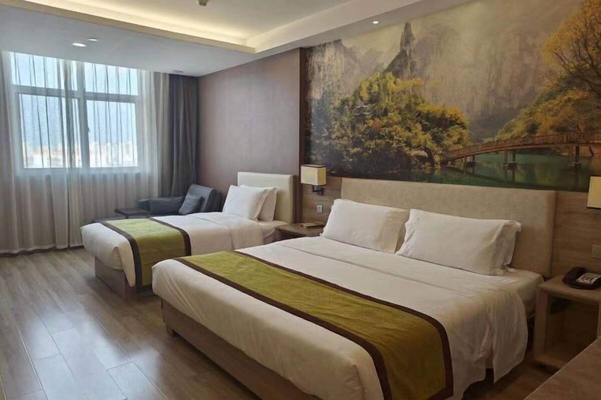 Ein Hotelzimmer mit zwei Betten, einem Landschaftsbild, einem Fenster mit Vorhängen und einem Nachttisch mit Lampe.