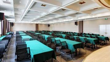 A conference room with rows of tables and chairs, a stage, and a white ceiling with recessed lighting.