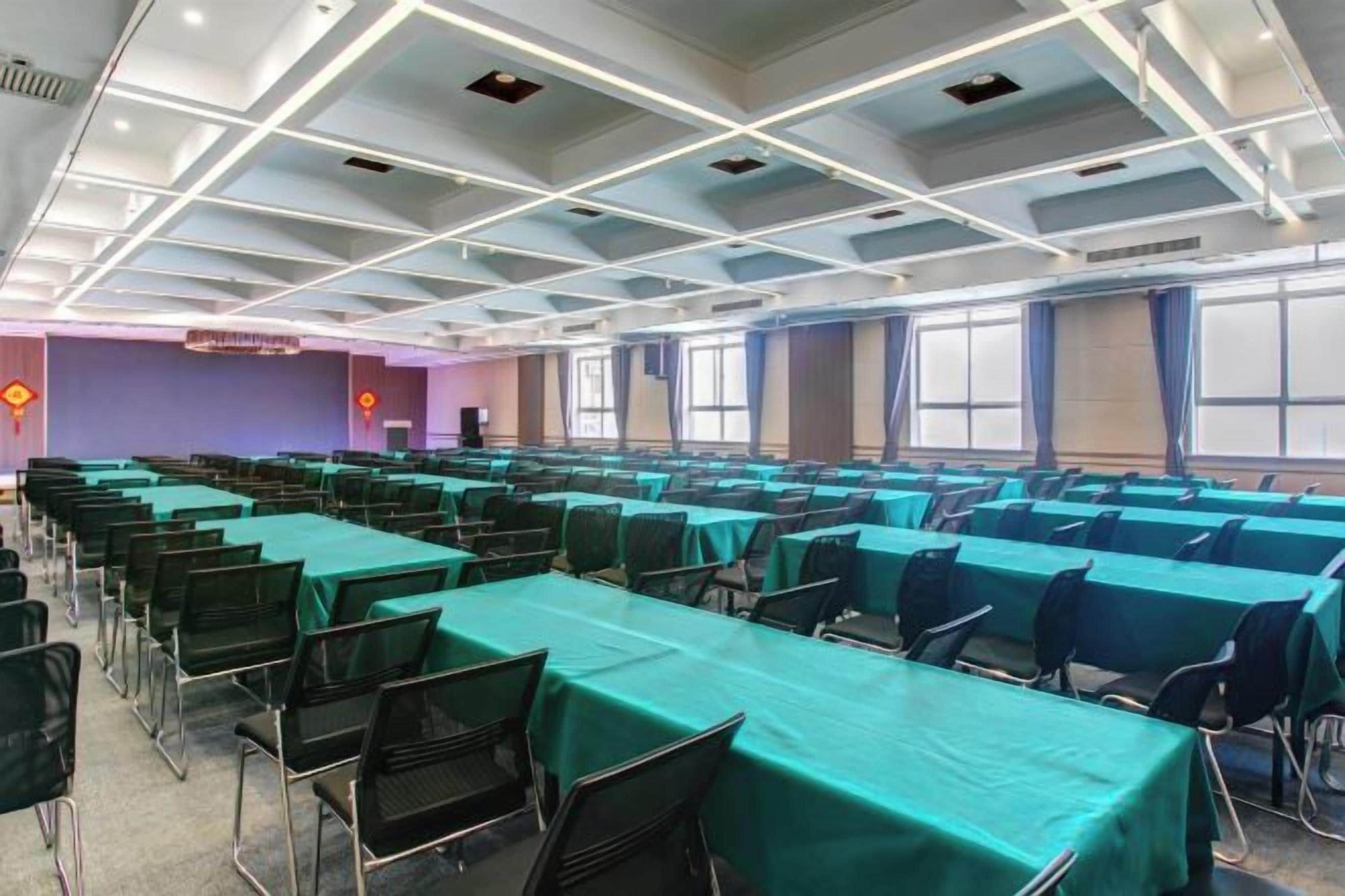 A conference room with rows of tables and chairs, large windows, and a high ceiling with lighting fixtures.