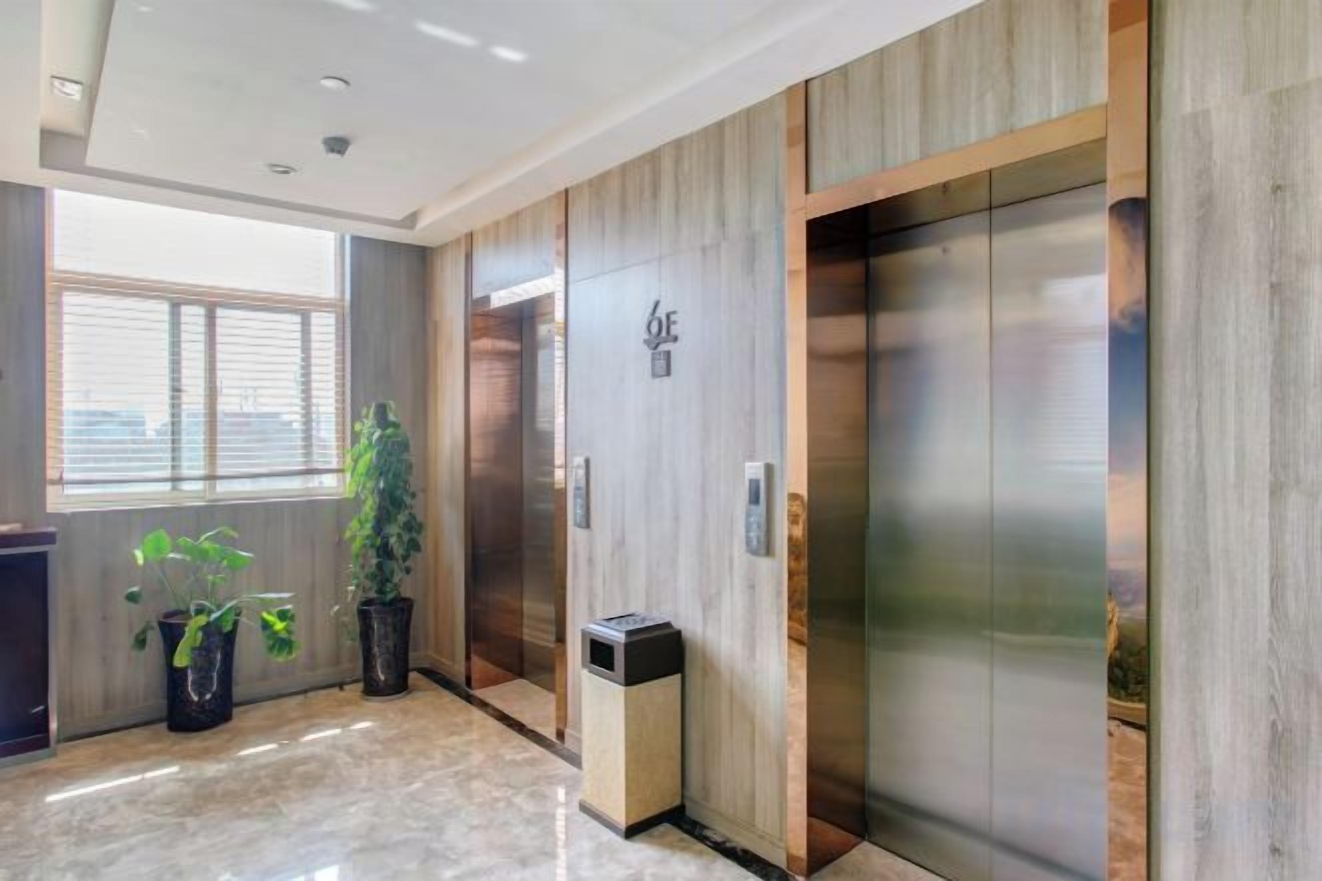 A hotel lobby with a wooden wall, an elevator, a trash bin, and potted plants.