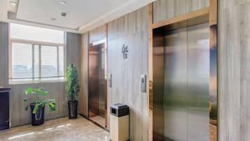 A hotel lobby with a wooden wall, an elevator, a trash bin, and potted plants.